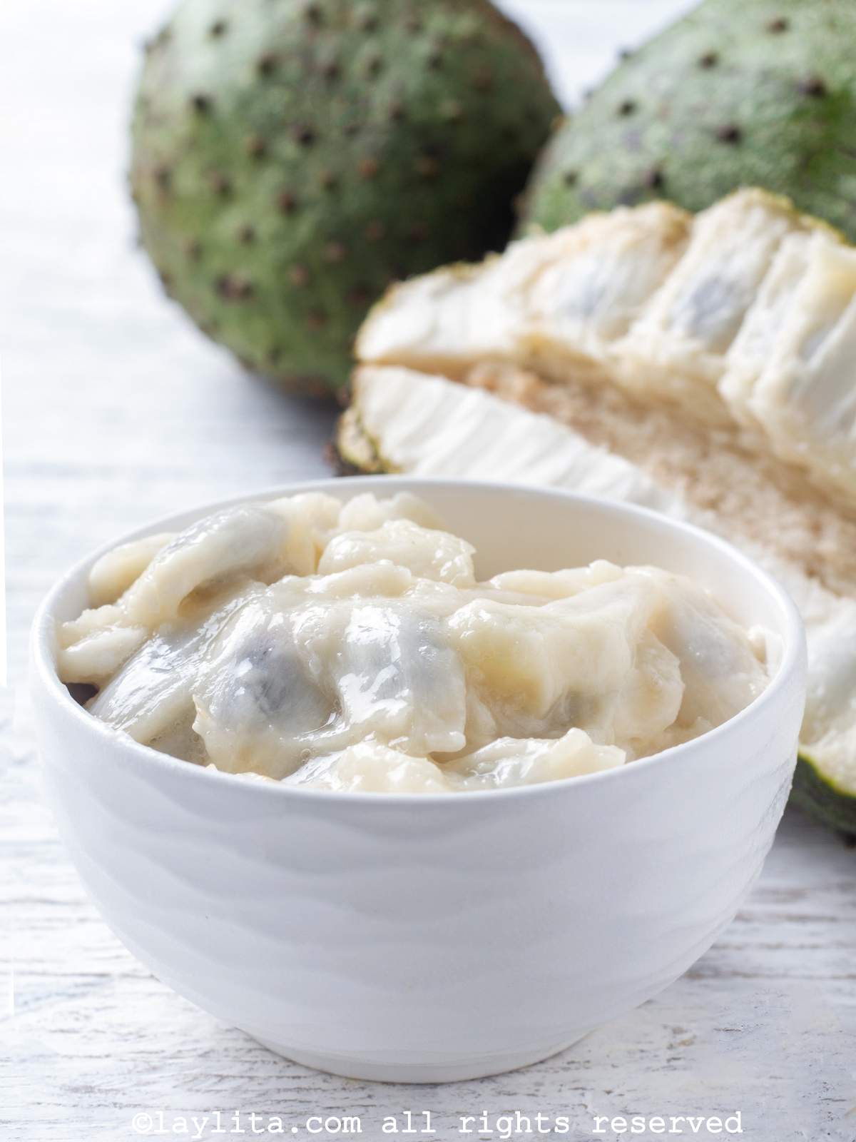 A bowl filled with guanabana or soursop fresh fruit pulp, with additional soursop fruits in the background