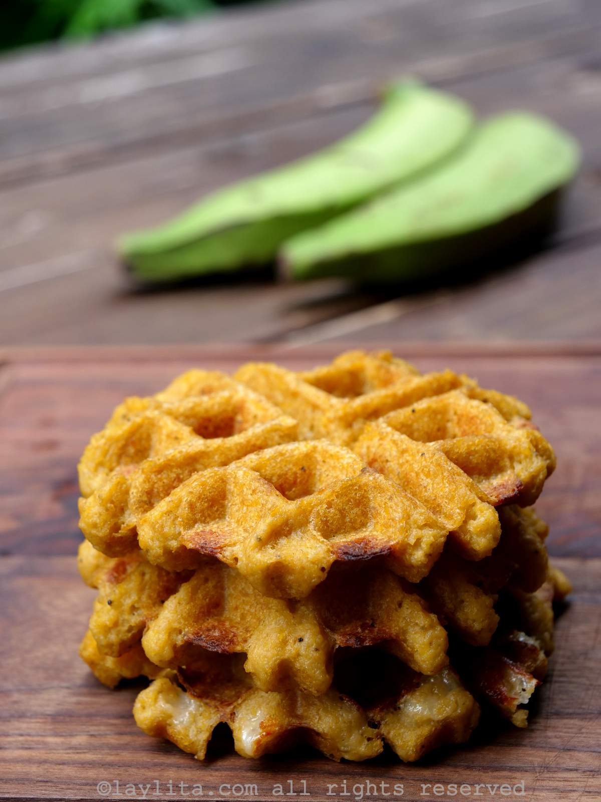 A stack of three green plantain waffles on a cutting board with two green plantains in the background
