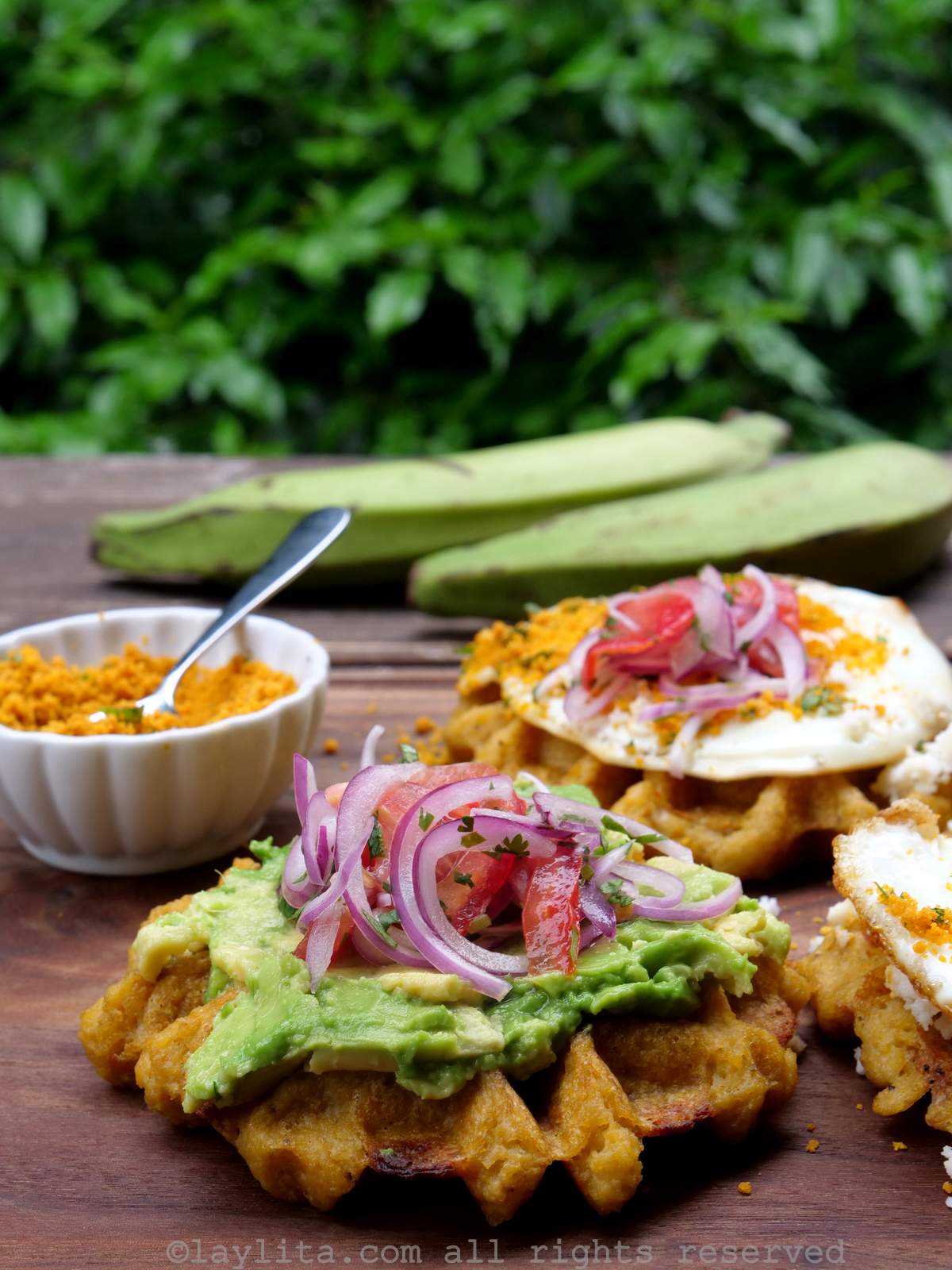A savory plantain waffle on a cutting board topped with mashed avocado, tomato and sliced onions, in the background there are more waffles and whole green plantains