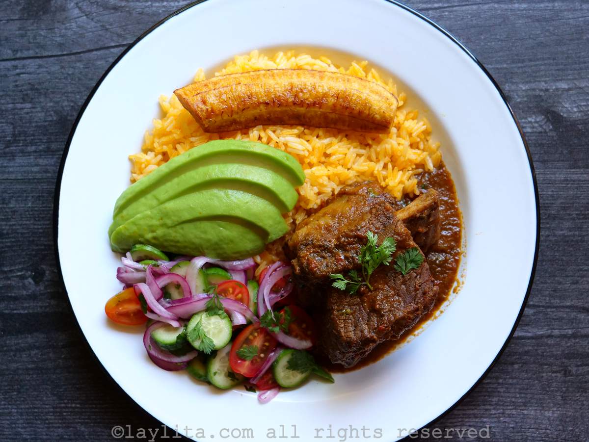 Seco de costilla beef rib stew, yellow rice, fried ripe plantains, avocado and a salad in a white plate with dark rim on top of a dark wooden table