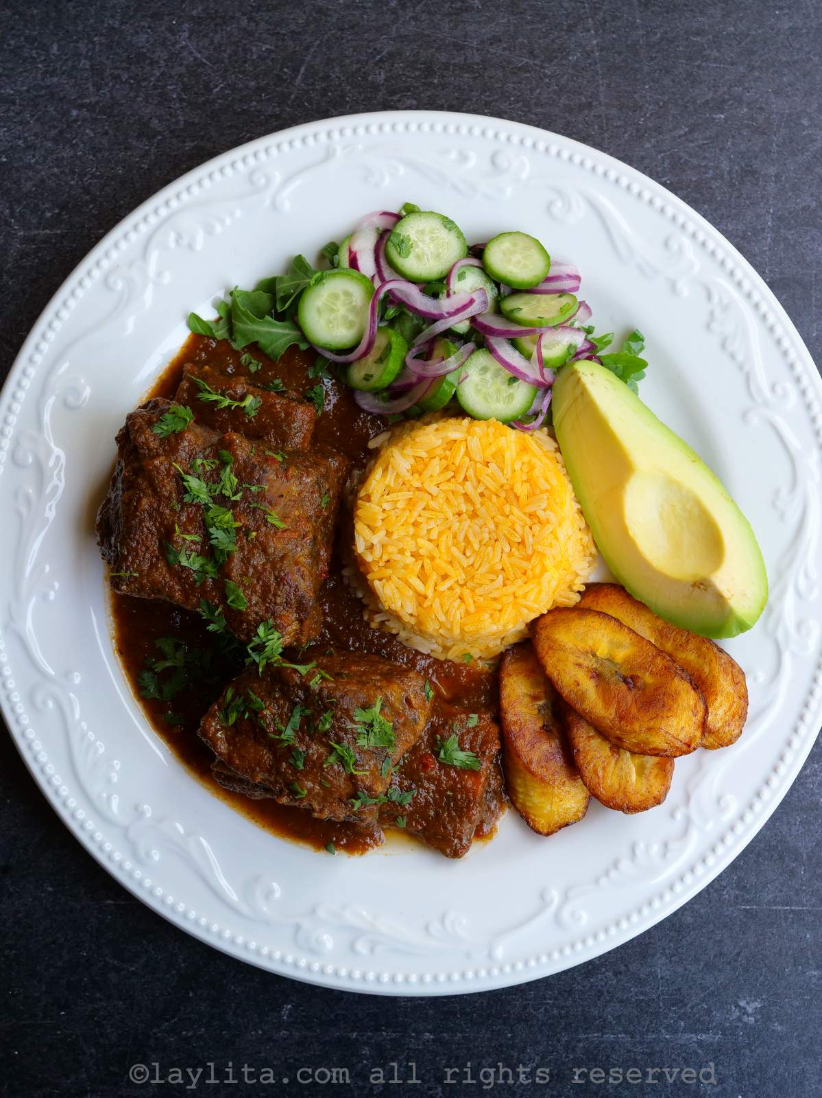 Overhead shot of a white plate with seco de costilla de res with sides like rice, avocado and cucumber salad.