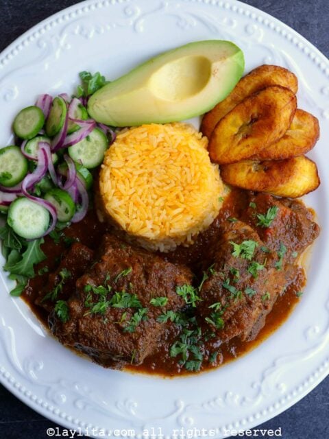 A white plate with seco de costilla beef rib stew, yellow rice, fried ripe plantains, avocado and a cucumber salad