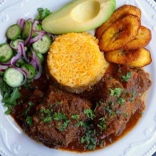 A white plate with seco de costilla beef rib stew, yellow rice, fried ripe plantains, avocado and a cucumber salad