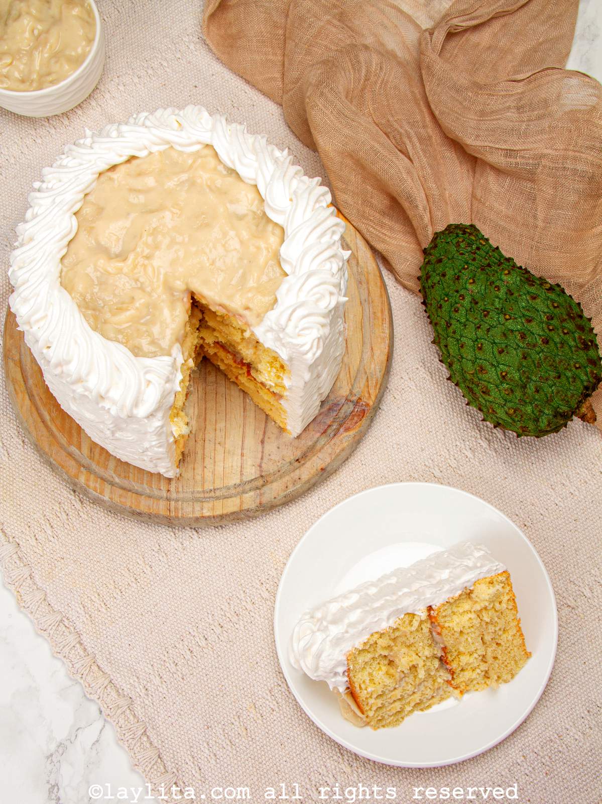 Overhead shot of a slice of soursop or guanabana cake on a white place with the rest of the cake behind it and a whole soursop fruit on the side