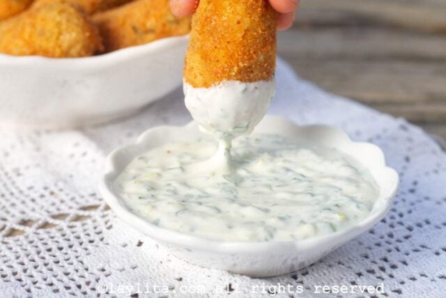 Fingers holding a crab croquette and dipping it into a white sauce