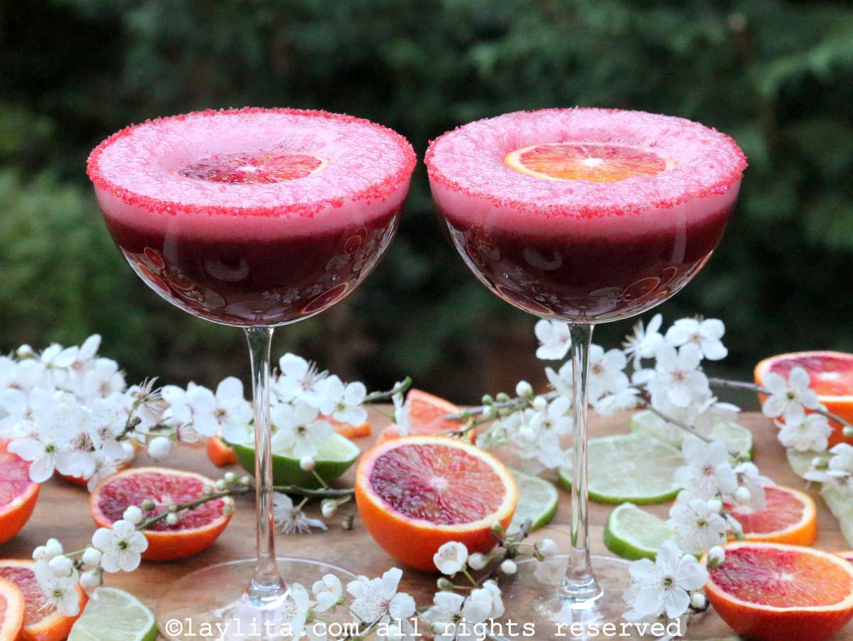 Two coupe glasses filled with blood orange pisco sours on a wooden board surrounded by blood orange fruit and flowers