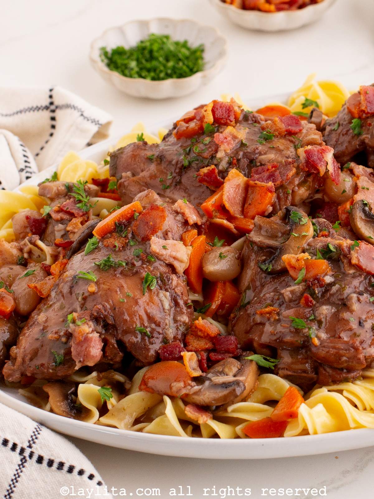 Chicken thighs braised in red wine over pasta on a large white platter with a bowl of chopped parsley in the background