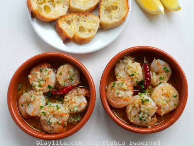 Overhead shot of two terracotta bowls of gambas al ajillo next to each other, with some bread slices and lemon wedges in the background