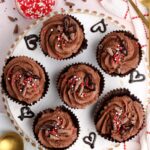 Overhead shot of six eggless chocolate mousse cups decorated with sprinkles and chocolate hearts on a wooden plate, with gold spoons and additional sprinkles on the side
