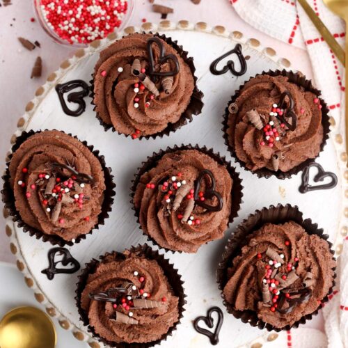 Overhead shot of six eggless chocolate mousse cups decorated with sprinkles and chocolate hearts on a wooden plate, with gold spoons and additional sprinkles on the side