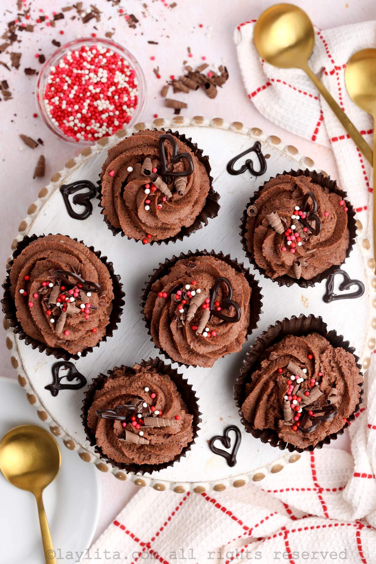 Overhead shot of six eggless chocolate mousse cups decorated with sprinkles and chocolate hearts on a wooden plate, with gold spoons and additional sprinkles on the side