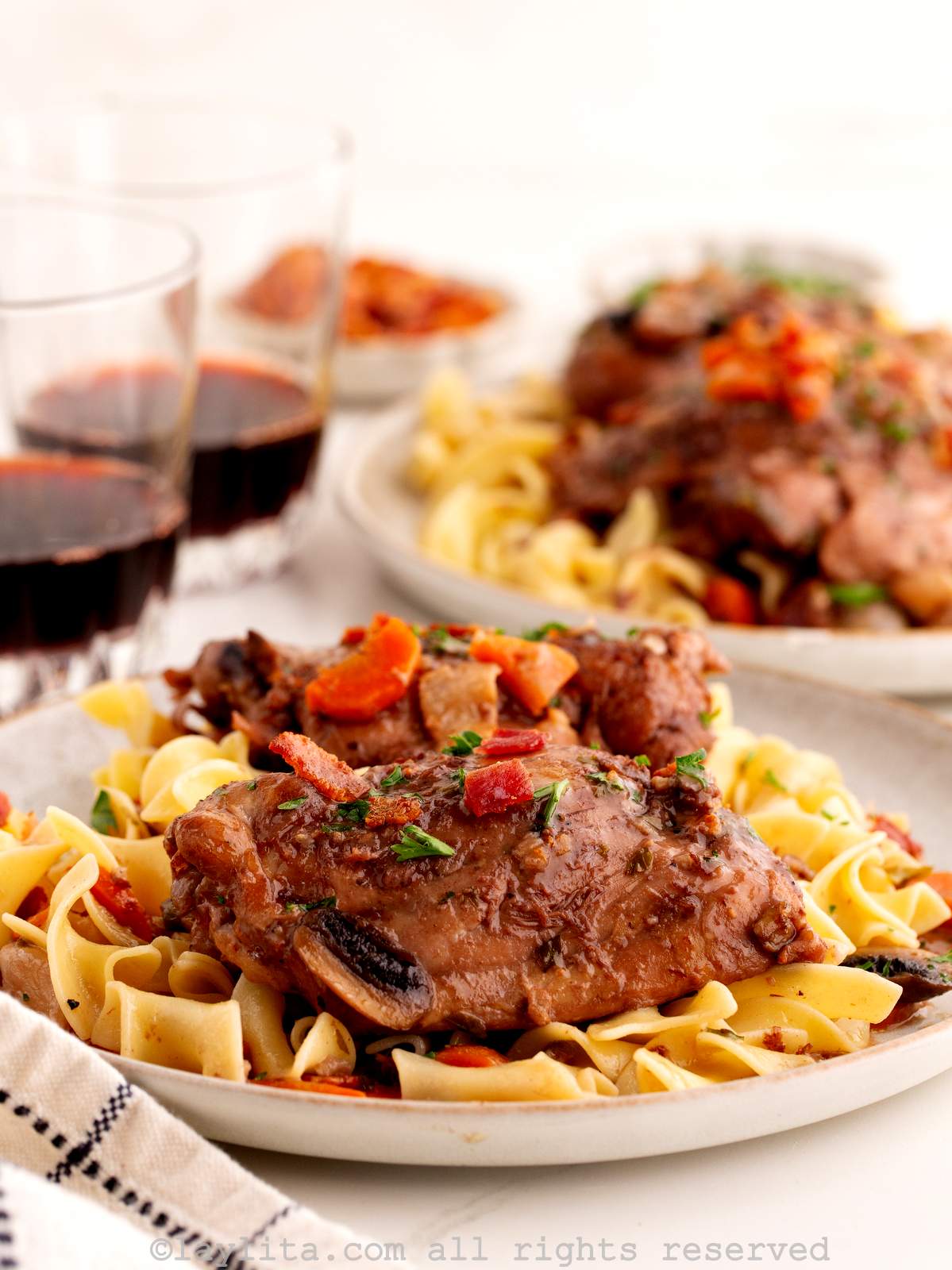Coq au vin chicken stew over pasta a white plate, with another plate and two glasses of red wine in the background