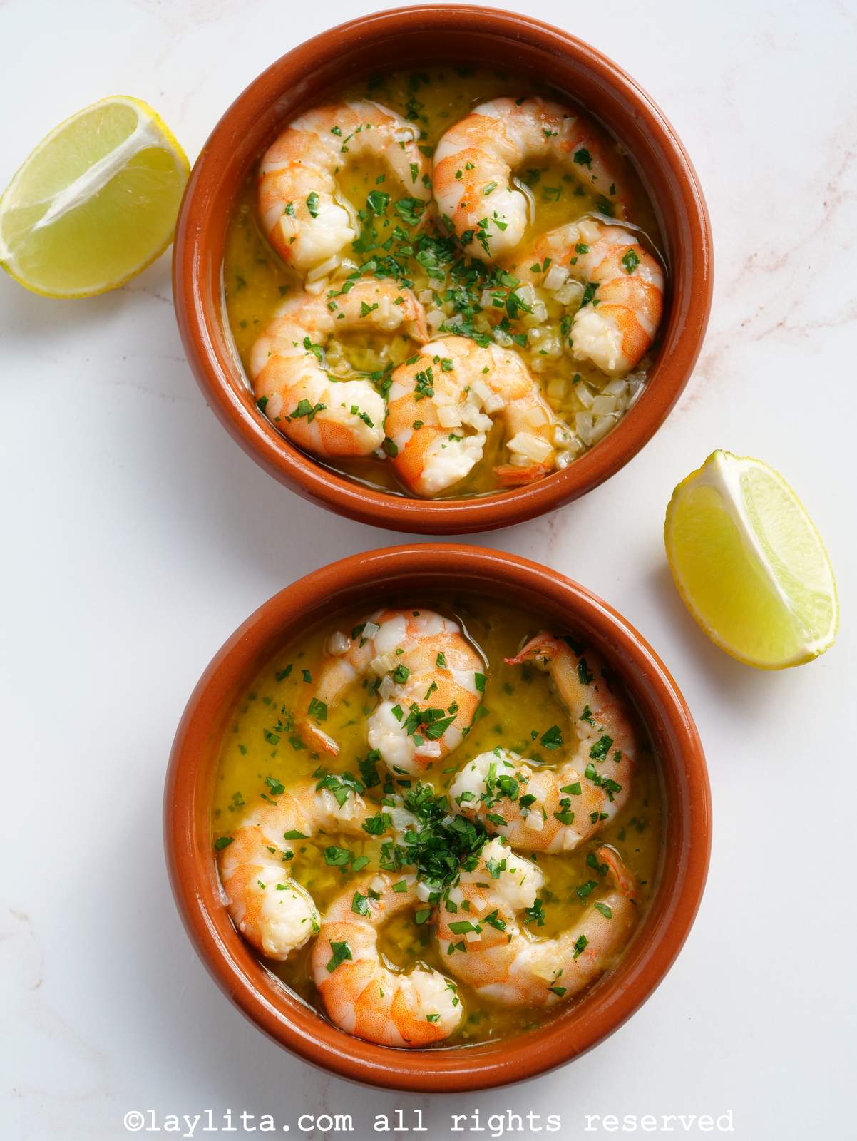 Two bowls of gambas al ajillo in a vertical line, with two lemon wedges next to them.
