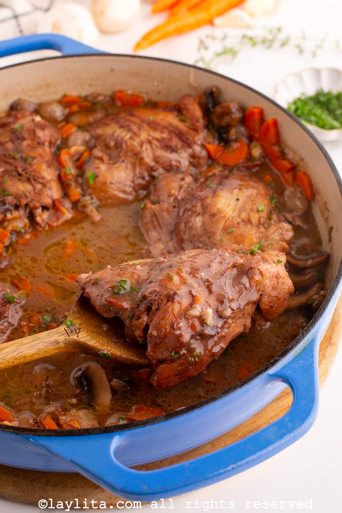 A blue stovetop filled with Coq au vin or French chicken cooked in red wine sauce, with a wooden spoon holding up a piece of chicken