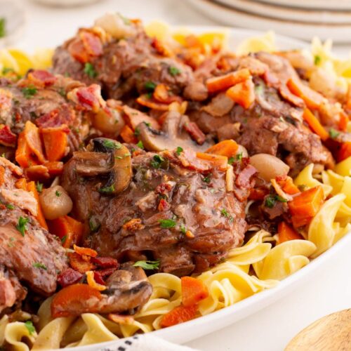 Side view of a platter filled with chicken thighs in red wine sauce served over egg noodles