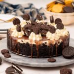 Oreo cheesecake topped with chocolate sauce, whipped cream and extra cookies, on a platter on a kitchen counter