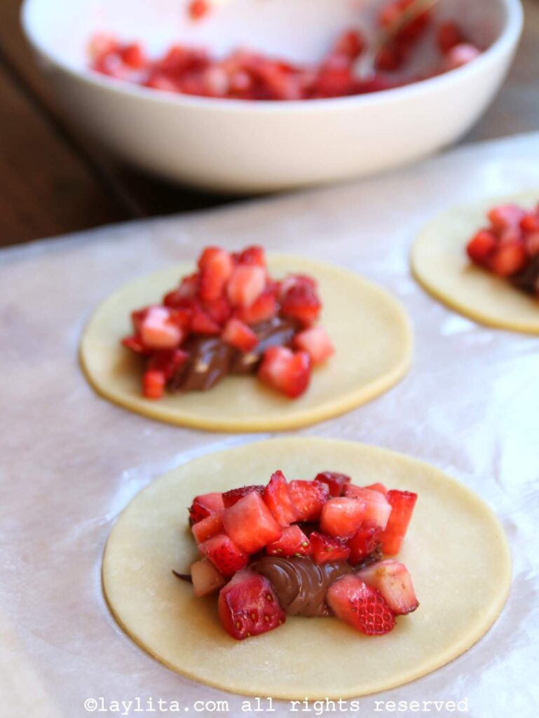 Filling the empanada dough with strawberries and Nutella on a wooden board with a bowl of diced strawberries in the background