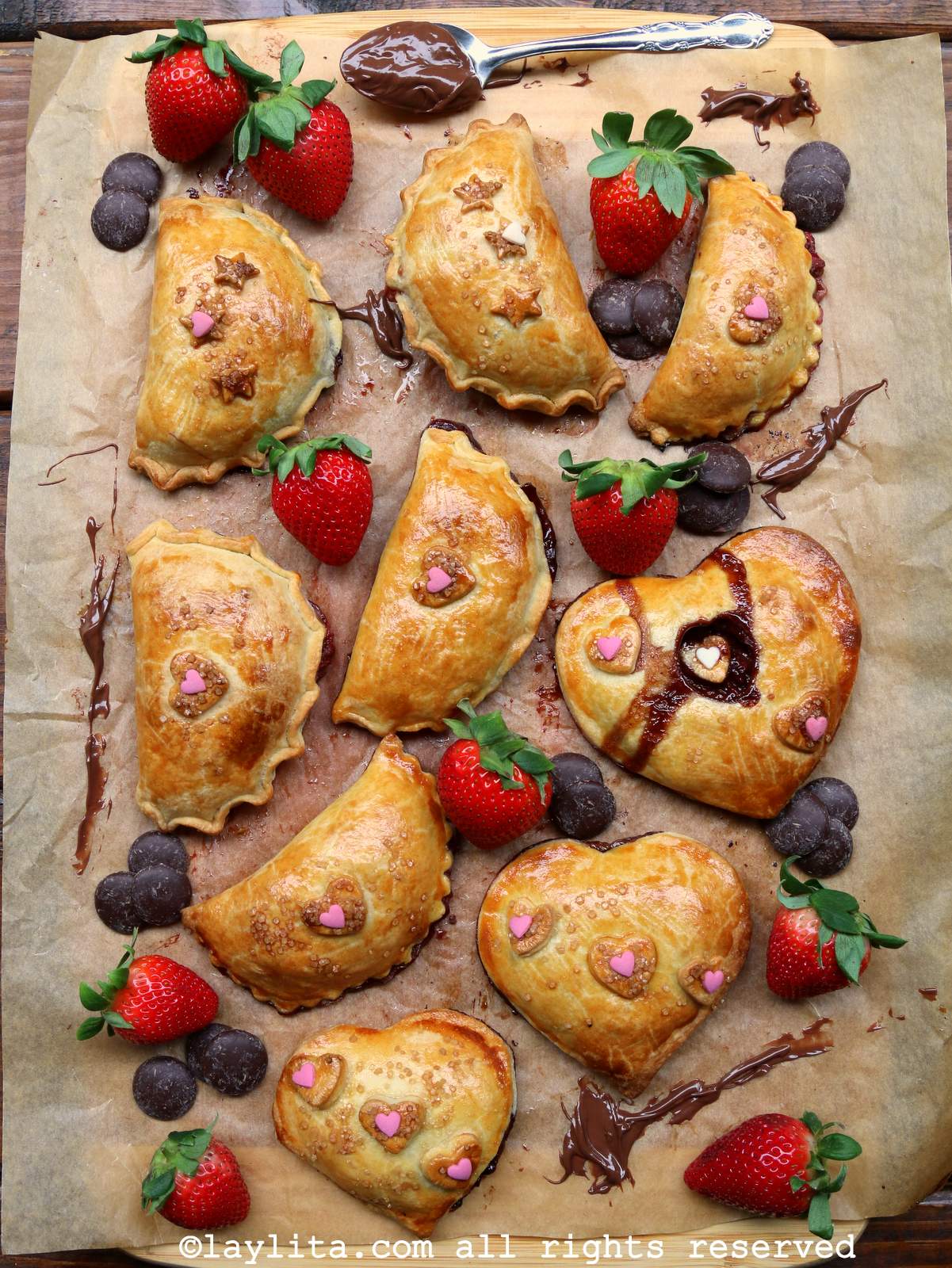Overhead shot of baked strawberry chocolate empanadas on a sheet pan lined with parchment paper, with fruit and chocolate chips scattered around them.