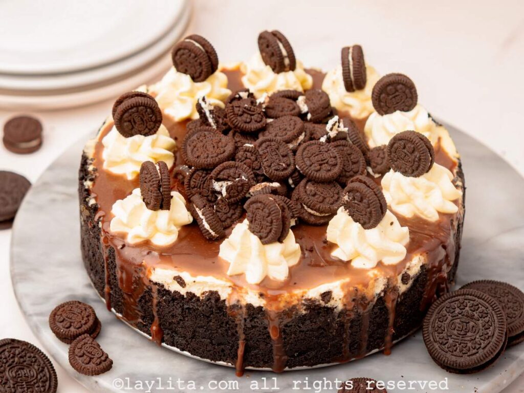 An Oreo cheesecake on a platter, the cheesecake is topped with chocolate sauce, whipped cream and mini oreos, with some additional cookies on the sides