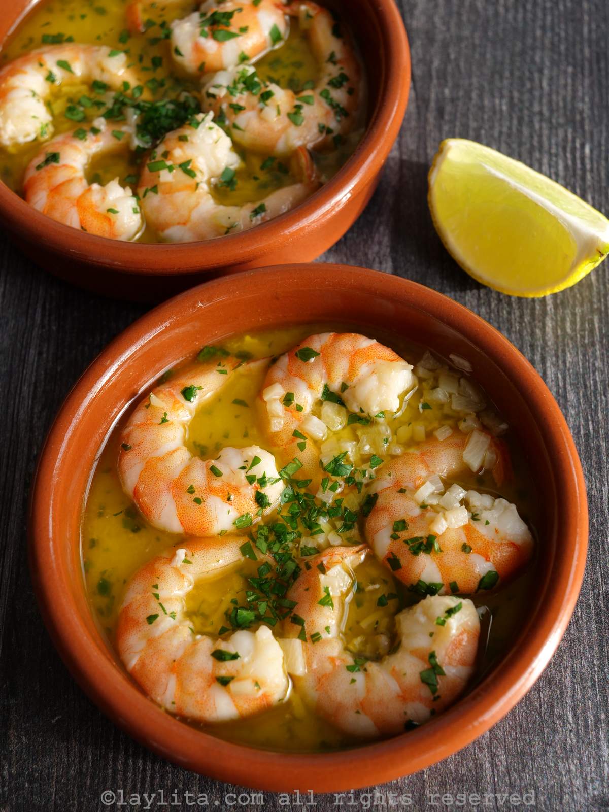 Two bowls of gambas al ajillo garnished with parsley, next to a lemon wedge.