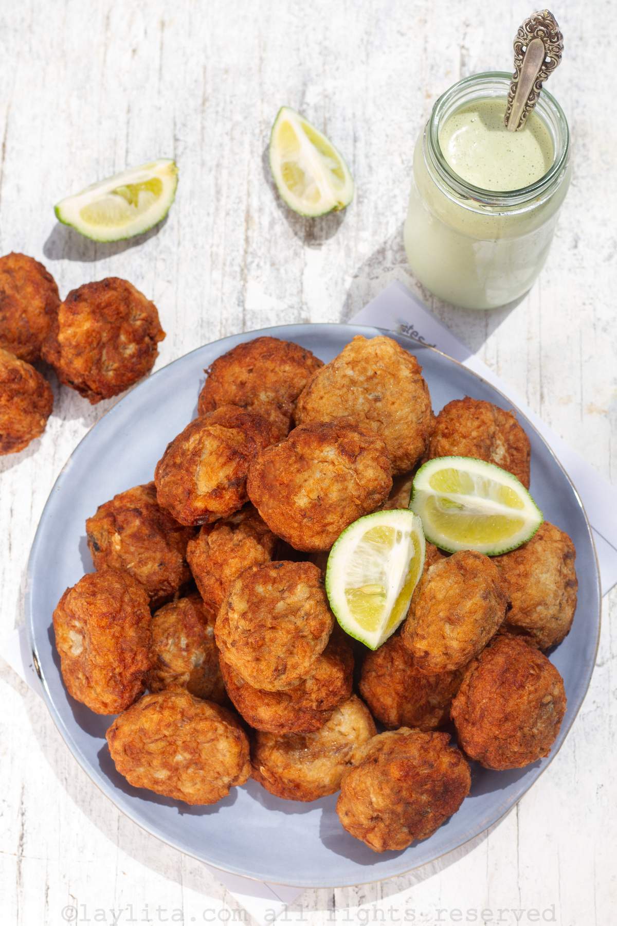 Overhead shot of salted cod fritters on a plate with lime wedges and a jar of green sauce next to it.