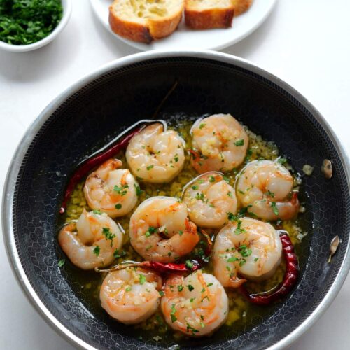 Overhead shot of a small black skillet filled with Spanish gambas al ajillo, with bread slices, chopped parsley and lemon wedges in the background