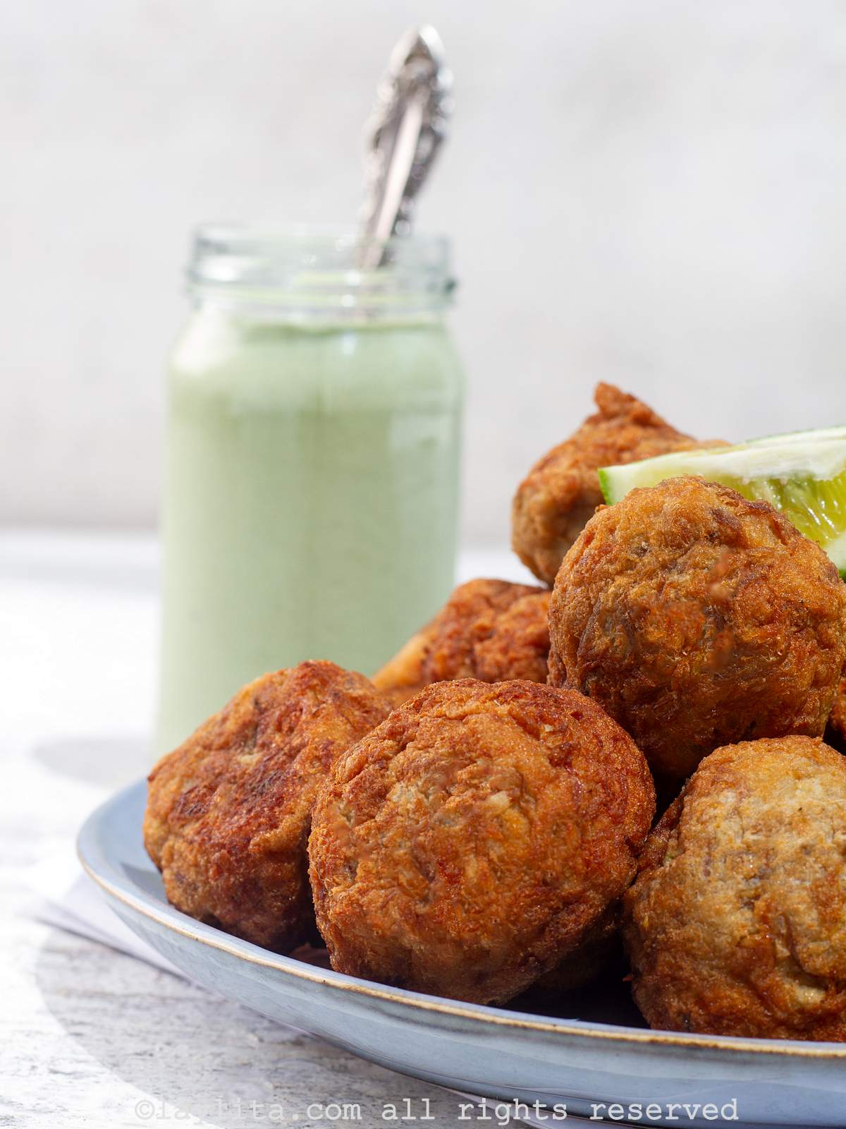 Salt cod fritters or buñuelos served on a plate with lime wedges with a green sauce behind it.