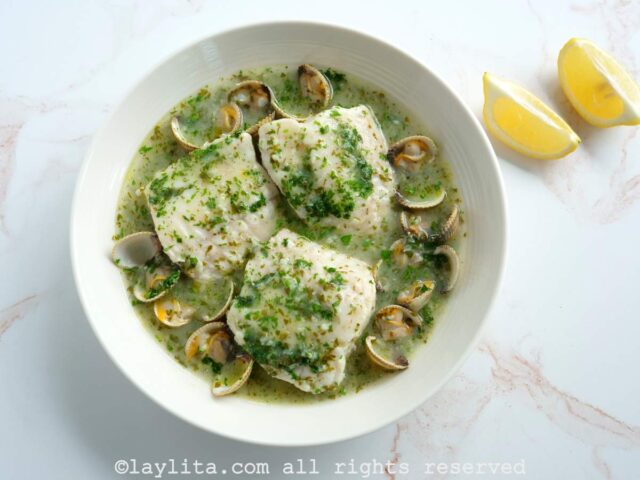 A white bowl filled with fish and clams in salsa verde on a marble countertop with a couple of lemon wedges on the side