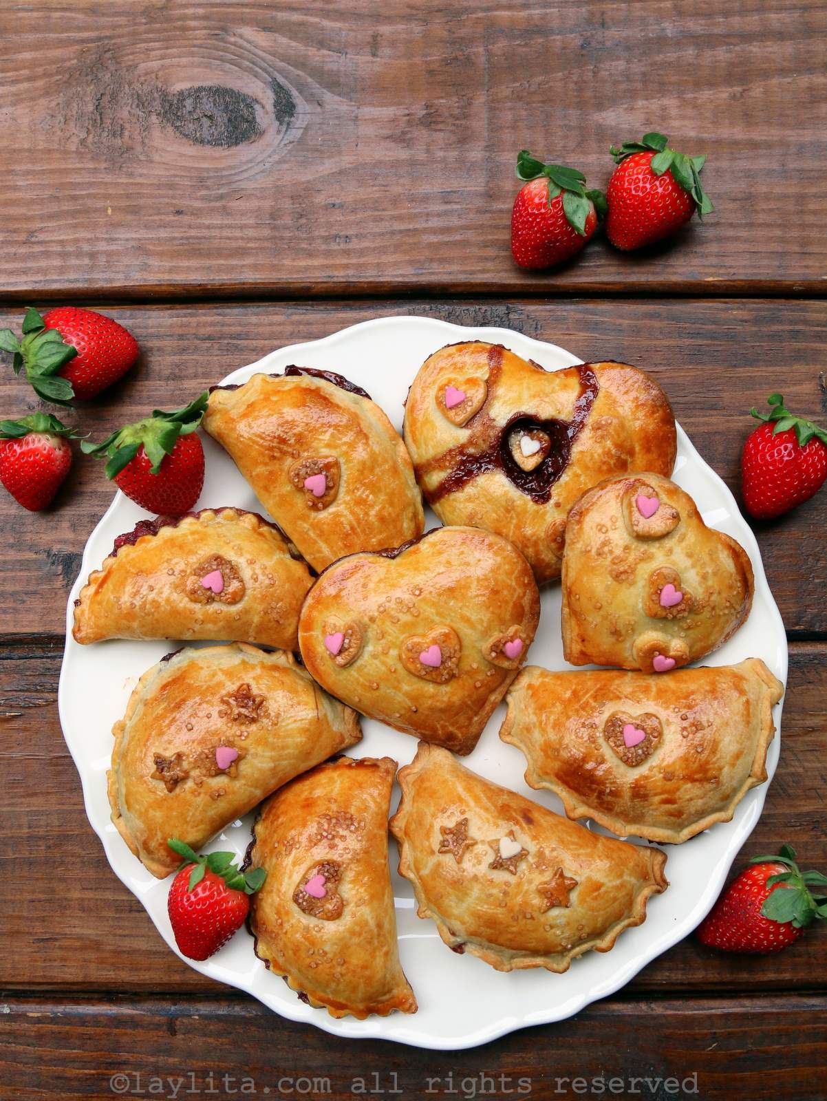 Overhead shot of strawberry chocolate empanadas on a white plate, some in half moon shapes and some in heart shapes on a wooden background.