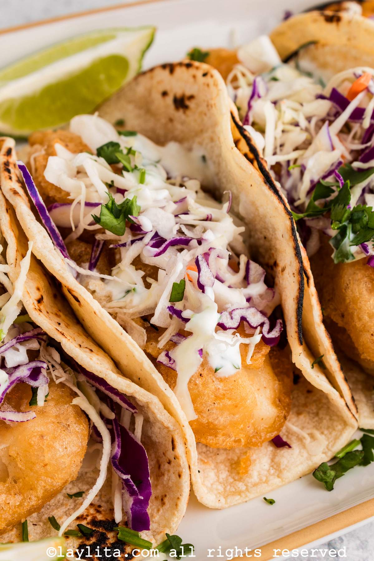 Closeup of a cod fish taco stacked next to other tacos, with a lime wedge in the back