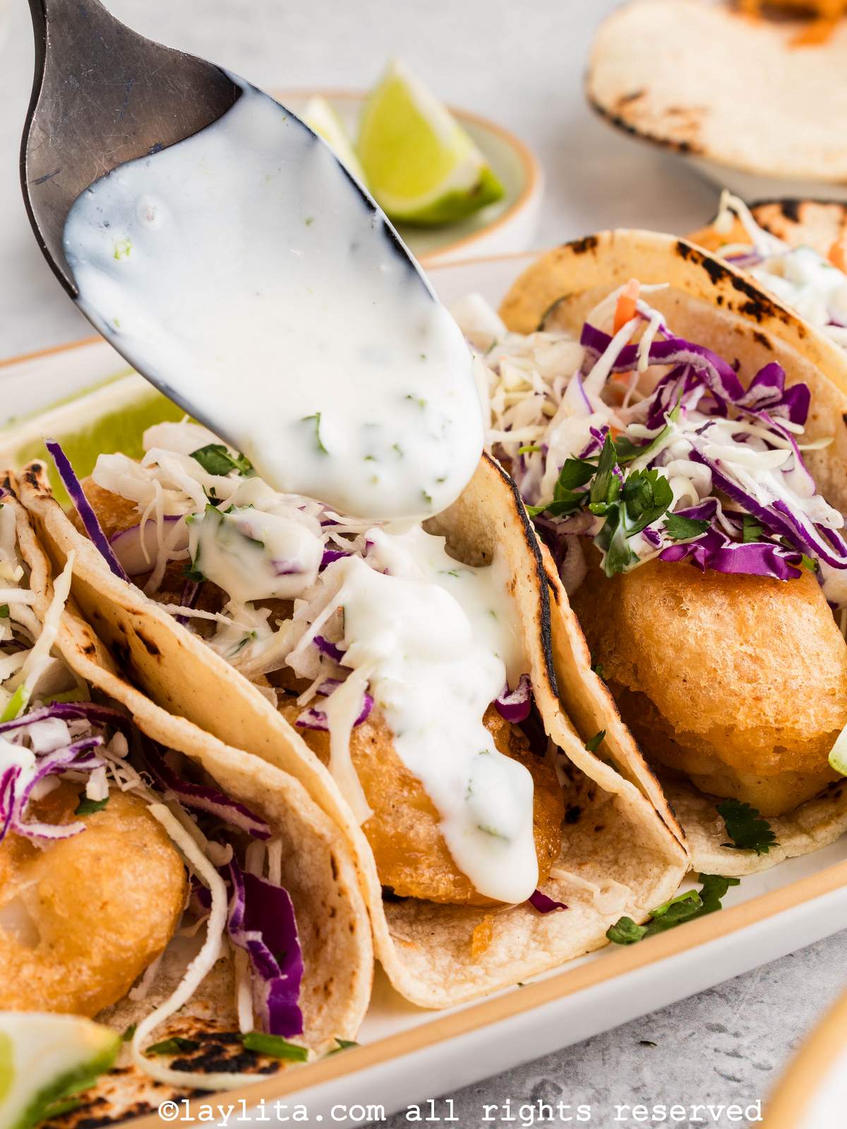 A spoon drizzling a creamy white sauce on top of a fish taco