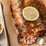 A large roasted salmon fillet on a baking sheet, there is a lemon sliced on top of the fish and more on the side