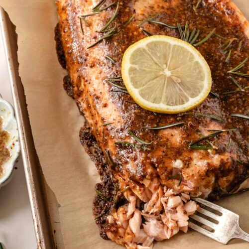 A large roasted salmon fillet on a baking sheet, there is a lemon sliced on top of the fish and more on the side