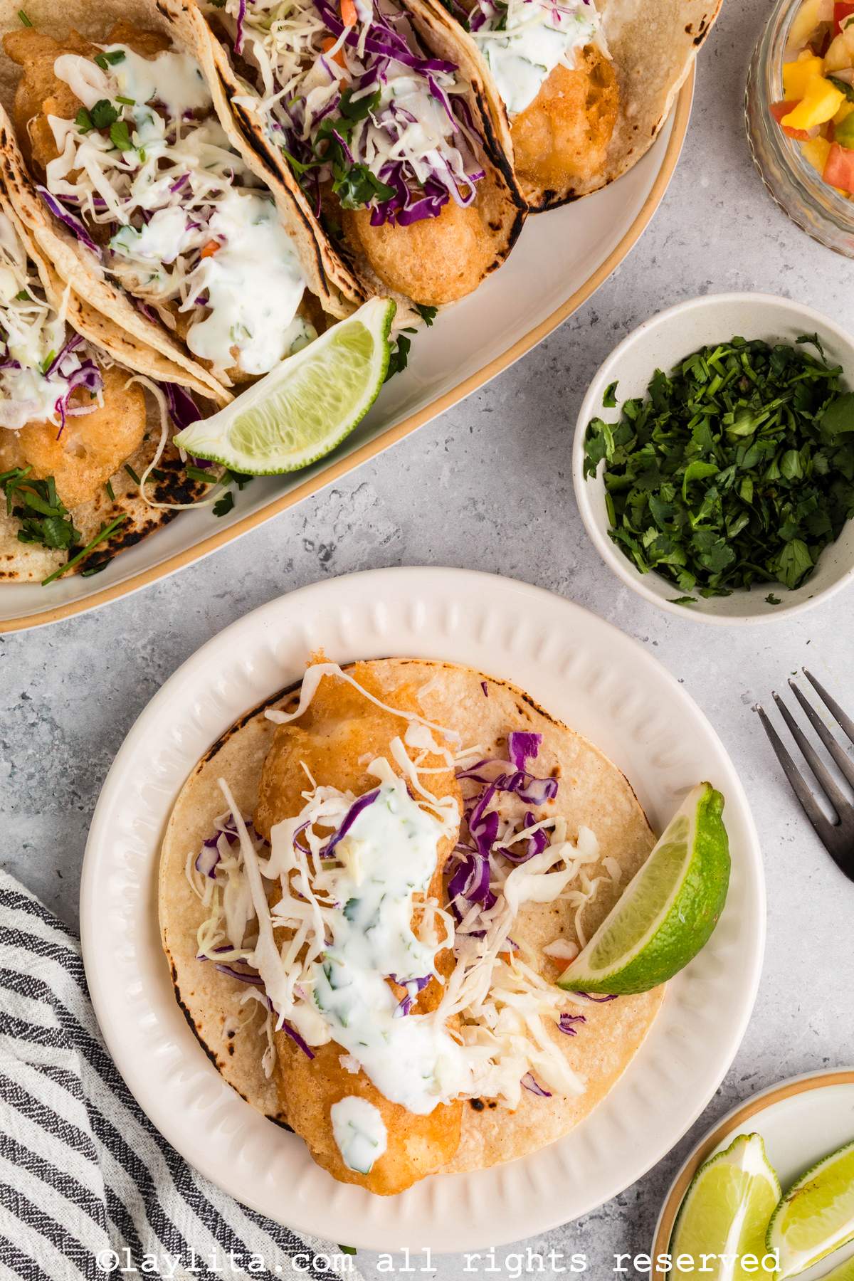 A Baja Fish taco on a white plate topped with lime crema sauce, with a large platter of tacos in the background
