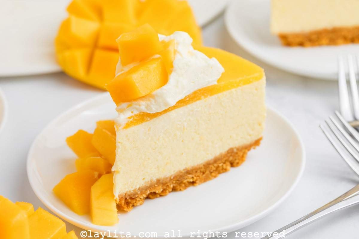Slice of mango cheesecake on a white plate garnished with whipped cream and mango chunks.
