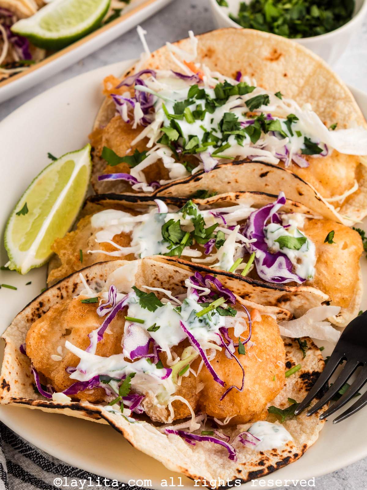 Three fish tacos on a plate, topped with mixed shredded cabbage and a creamy white sauce, with a lime wedge on the side