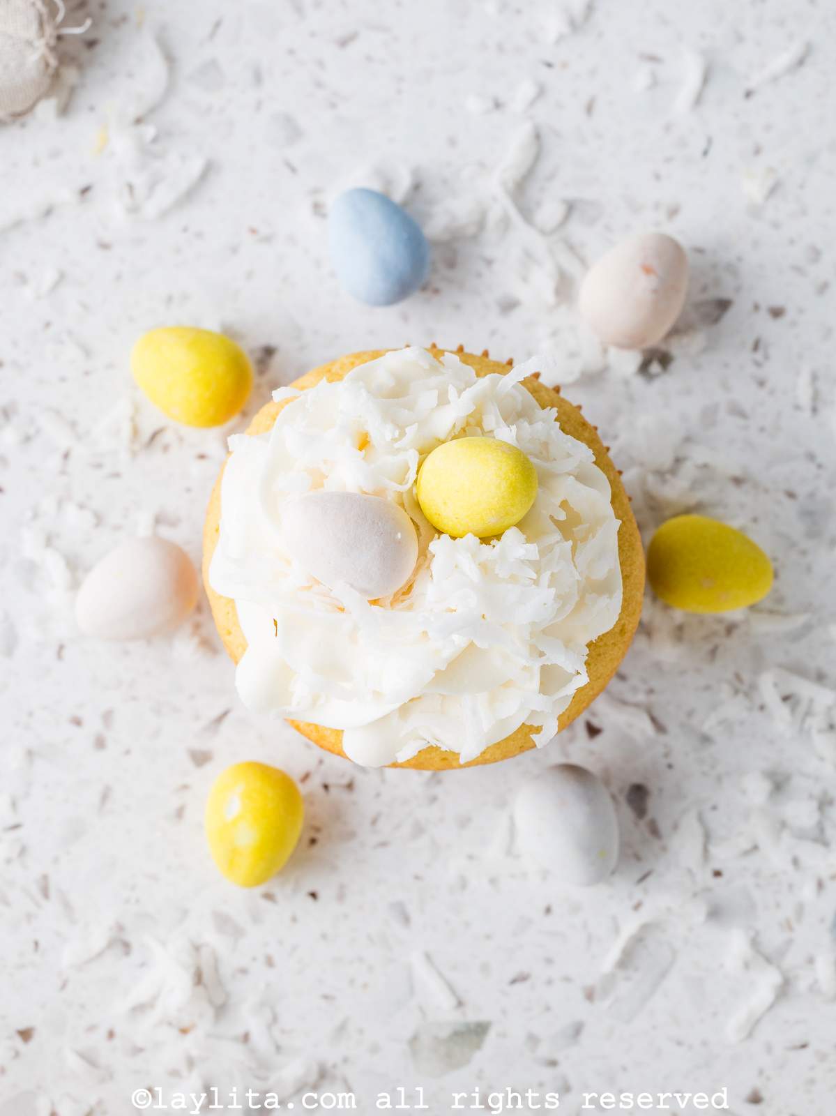 Overhead view of a coconut cupcake topped with frosting and mini-egg candies, with more candies around it