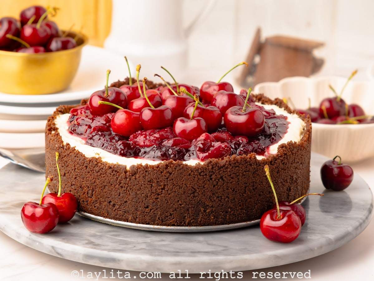 A cherry cheesecake with a chocolate cookie crust on a marble platter with a few cherries on side