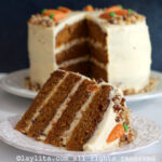 A slice of layered carrot cake on a white plate with the rest of the cake in the background