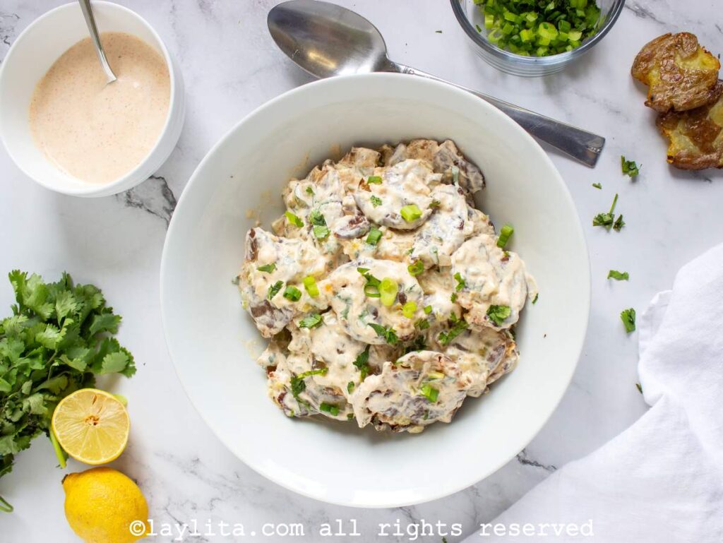 Overhead shot of smashed potato salad next to the dressing, herbs, and a lemon cut in half.