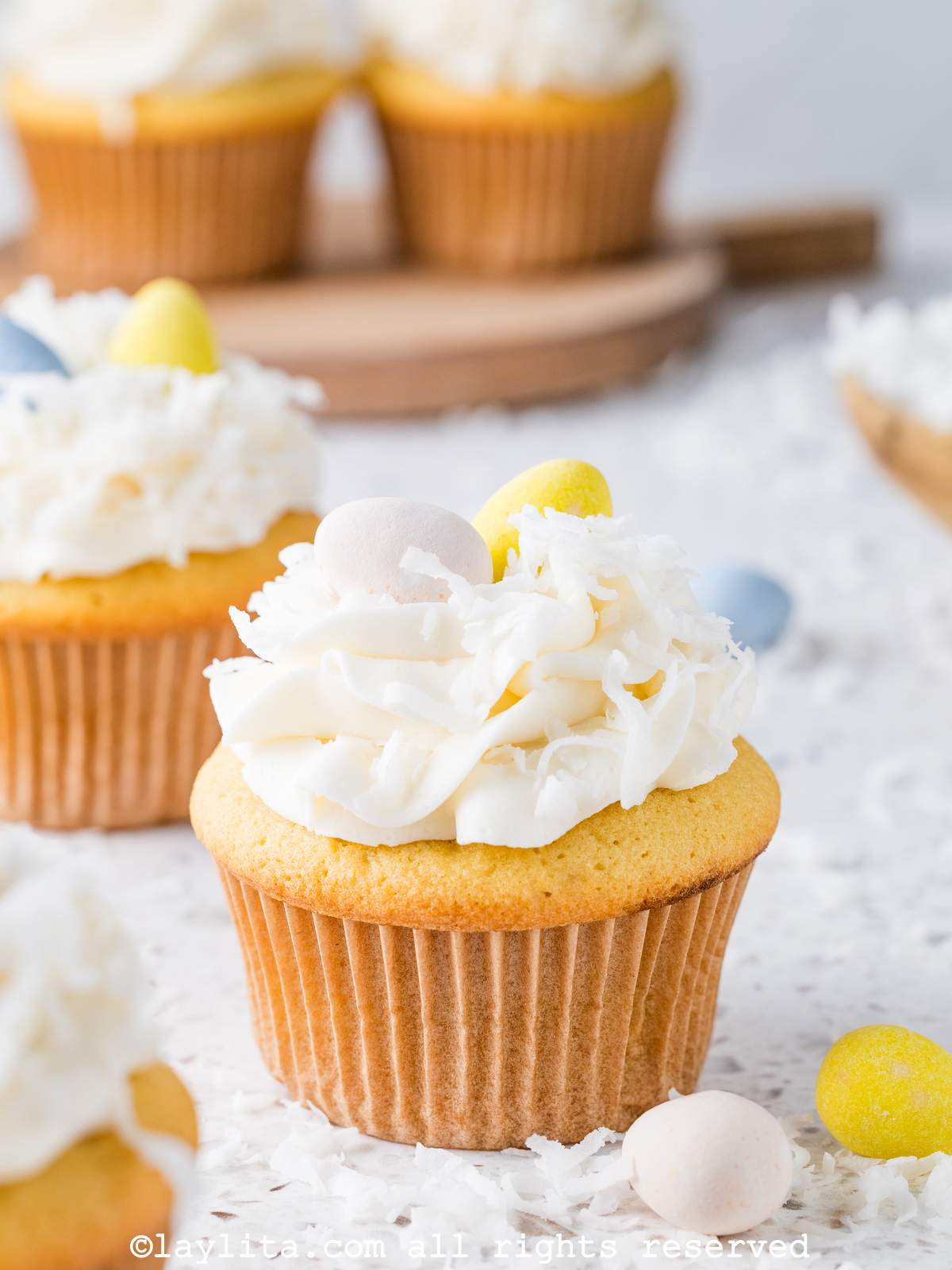 A coconut cupcake topped with coconut flakes and easter egg decorations on a kitchen counter with more cupcakes in the background
