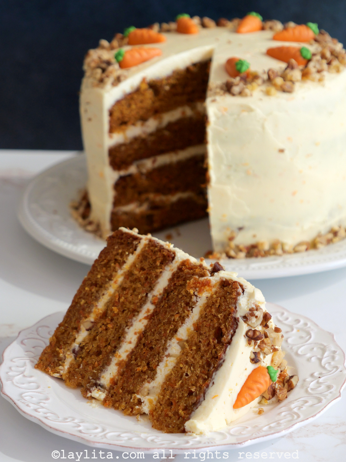 Closeup of a slice of layered carrot cake on a white plate with the rest of the cake in the background