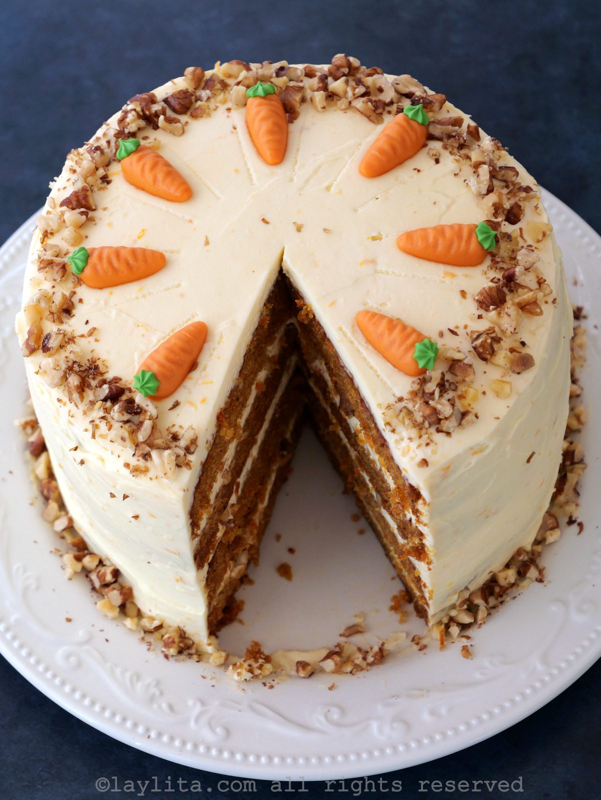 Overhead view of a layered carrot cake with a slice missing, showing the four layers, the cake is decorated with cream cheese, crushed nut and fondant carrots