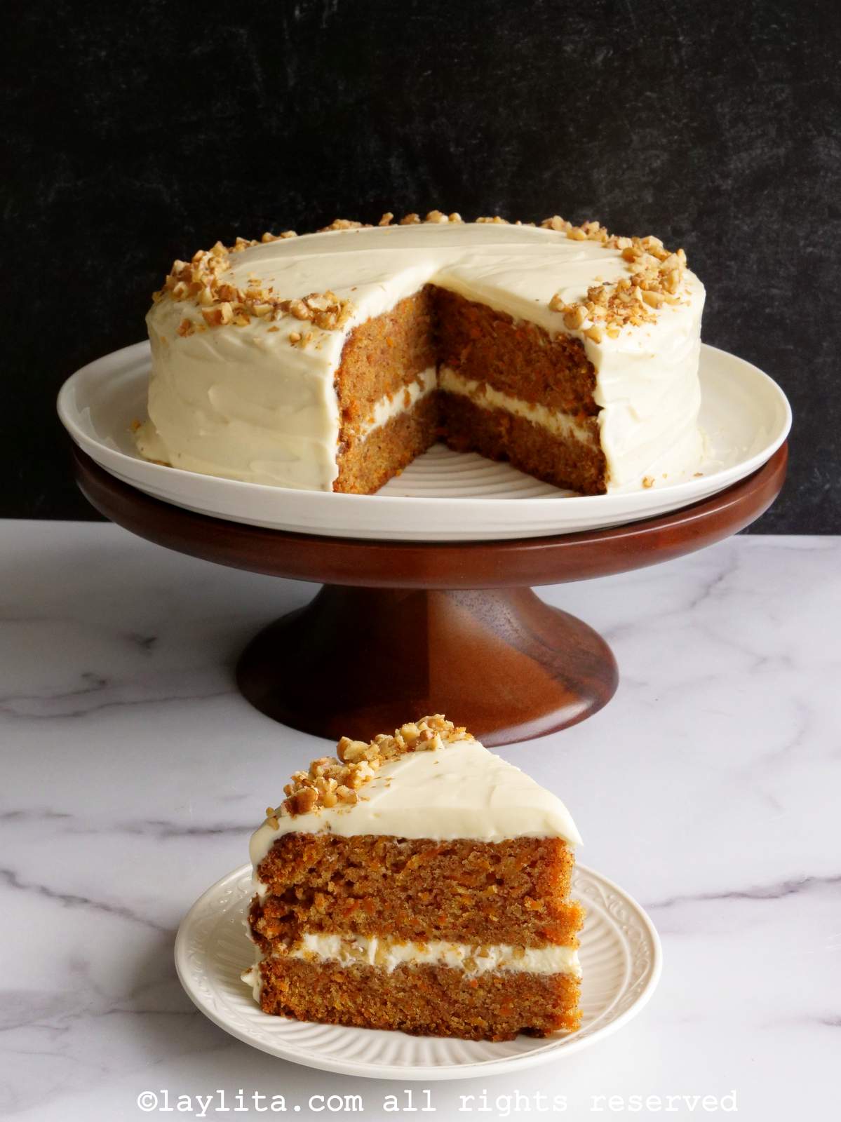 A slice of carrot cake with two layers on a plate, with the rest of the cake on a cake serving platter in the background