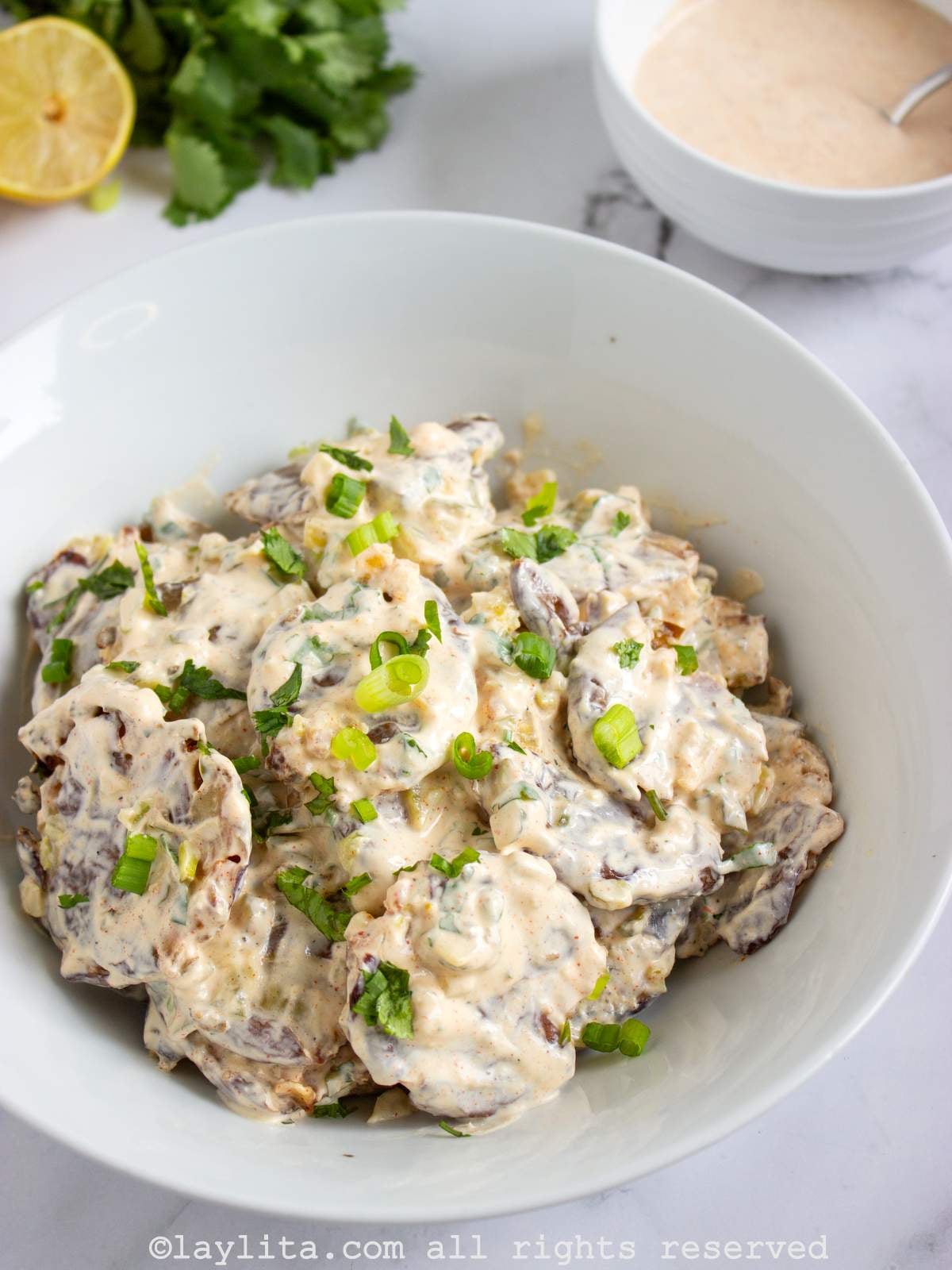 Smashed potato salad in a white bowl with green onions on top, there is a small bowl of creamy sauce and a lemon cut in half in the background