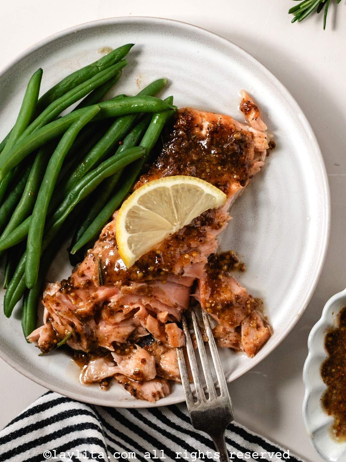 A piece of roasted maple mustard salmon on a white plate next to green beans with a lemon slice on top