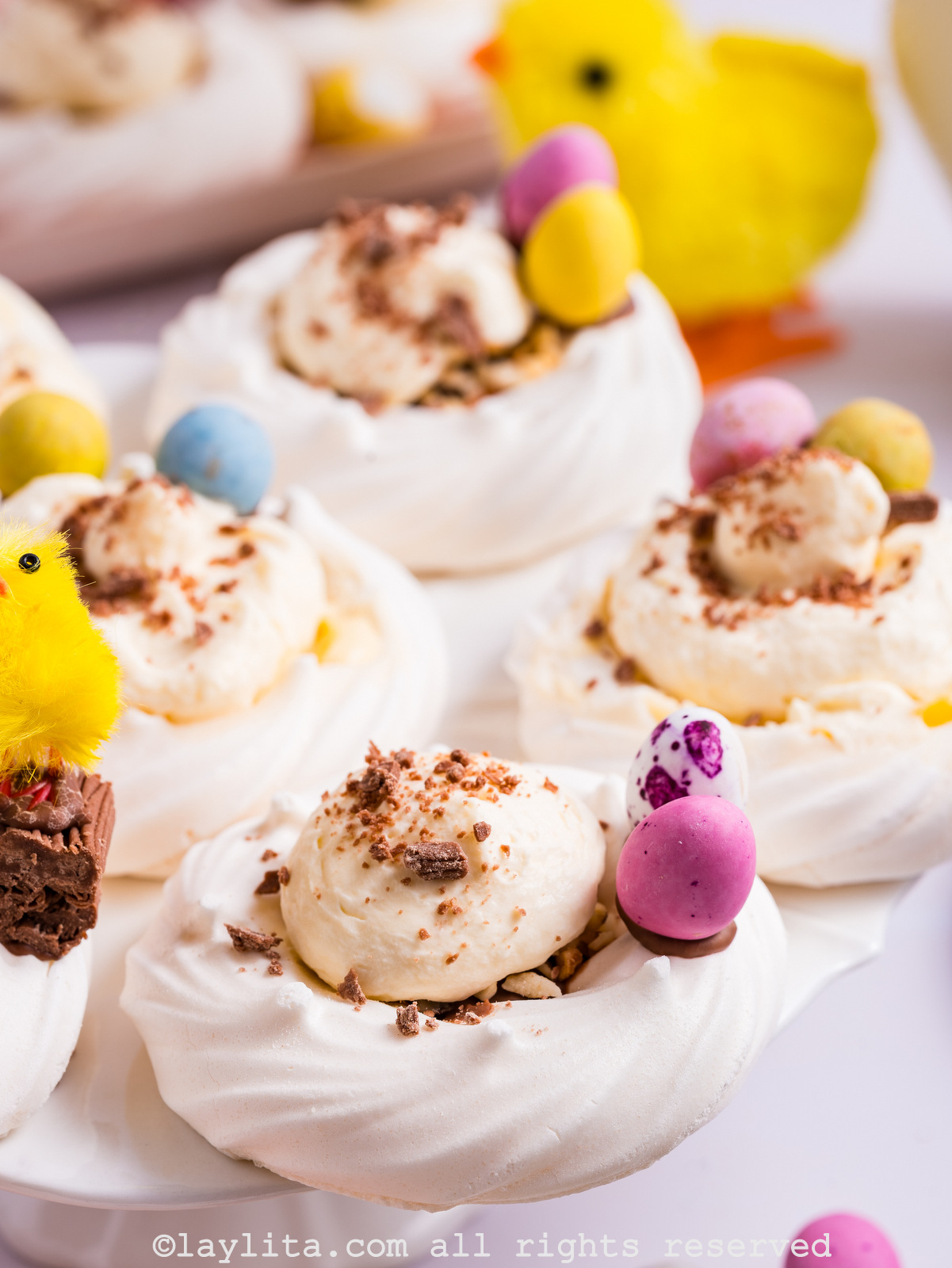 Several meringue nests filled with lemon custard cream and topped with chocolate shavings, with mini eggs on the side