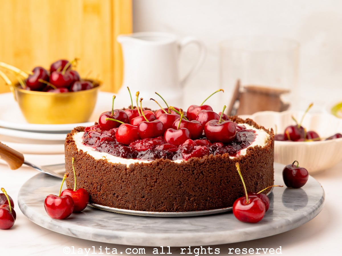 A no bake cherry cheesecake with a chocolate cookie crust on a marble platter with a few cherries on side and more in the backgroun