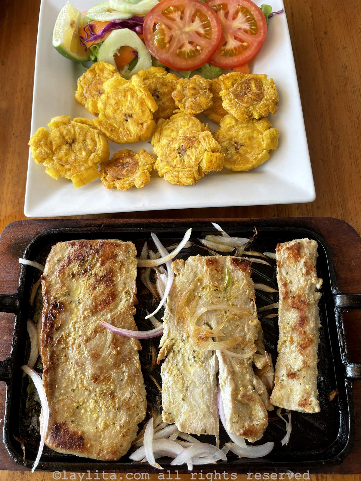 A plate of grilled tuna a la plancha with onions next to a plate with patacones and salad in Ecuador