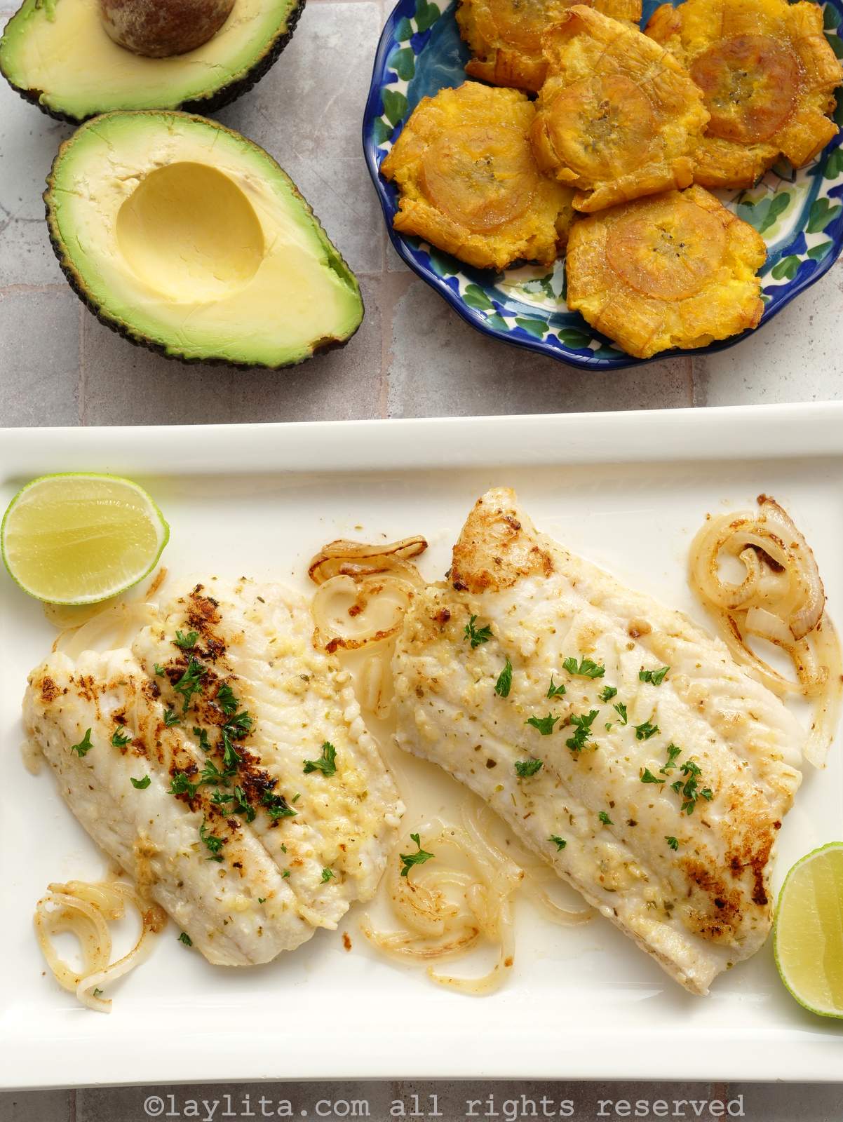 Grilled fish on a serving platter with lime and onions, in the background there is a plate with fried green plantains and an avocado cut into halves